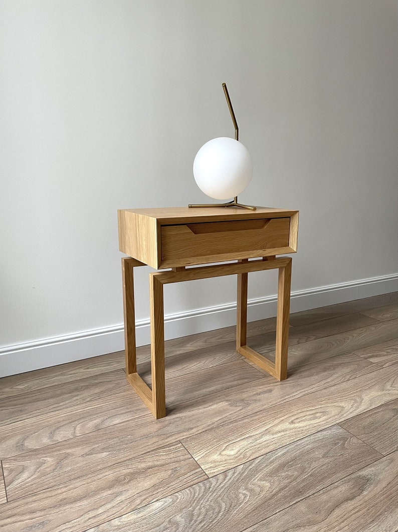 May include: A wooden nightstand with a single drawer and a metal lamp with a white globe. The nightstand has a simple, modern design and is made of light wood.