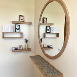 May include: A wooden wall shelf with a round mirror above it. The shelf is decorated with small framed pictures, books, and a vase with dried flowers. The mirror is oval shaped and has a wooden frame.