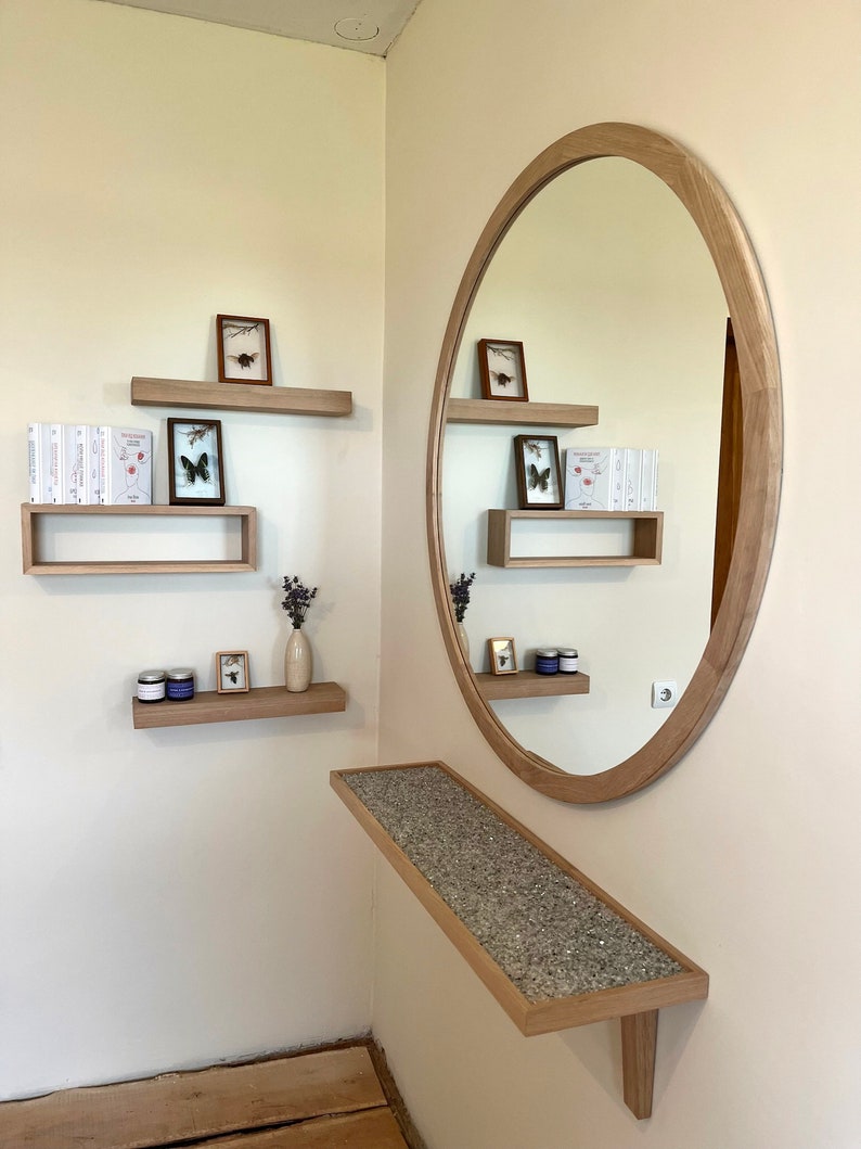 May include: A wooden wall shelf with a round mirror above it. The shelf is decorated with small framed pictures, books, and a vase with dried flowers. The mirror is oval shaped and has a wooden frame.