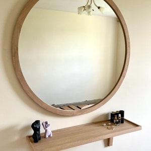 May include: A round wooden framed mirror with a shelf underneath. The shelf has two small decorative sculptures and a few bottles of perfume.