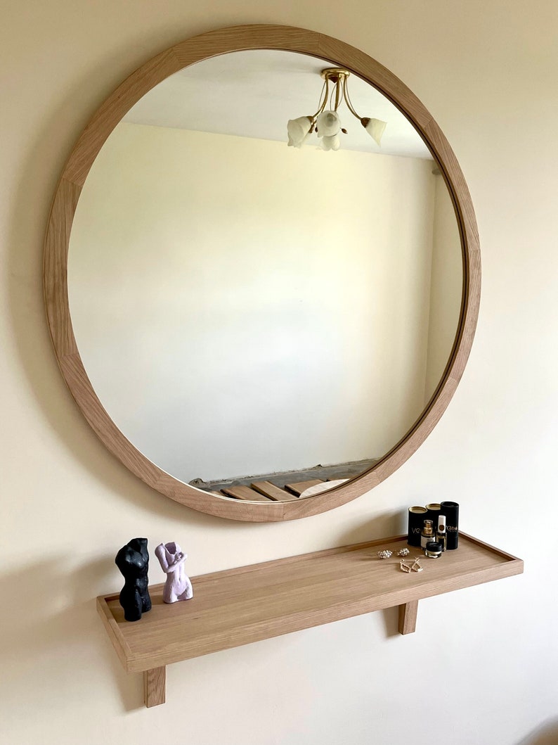 May include: A round wooden framed mirror with a shelf underneath. The shelf has two small decorative sculptures and a few bottles of perfume.