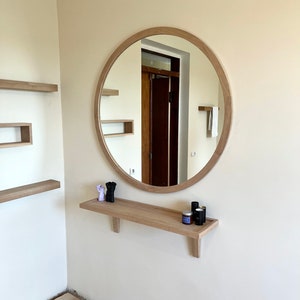 May include: A round wooden framed mirror with a shelf underneath. The shelf has two small purple figurines and three small bottles on it.