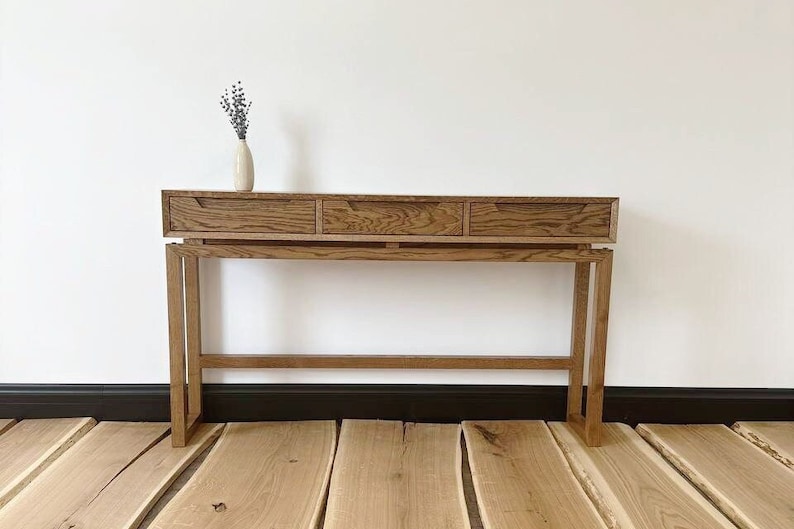 May include: A wooden console table with three drawers and a simple, modern design. The table is made of light brown wood and has a natural finish. It is sitting on a wooden floor.