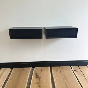 以下が含まれることがあります： 白い壁に取り付けられた、引き出し付きの黒いフローティングナイトスタンドが2つ。ナイトスタンドは木でできており、モダンなデザインです。