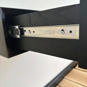 以下が含まれることがあります： 黒い引き出しのスライドのクローズアップ。スライドには「HAFELE」という文字が印刷されています。スライドは金属製で、銀色の仕上げになっています。