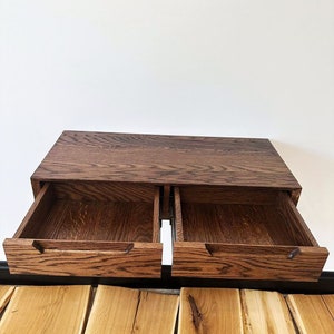 May include: A wooden wall-mounted shelf with two drawers that are open. The shelf is made of dark wood with a grain pattern. The drawers are also made of wood and have a light brown finish.