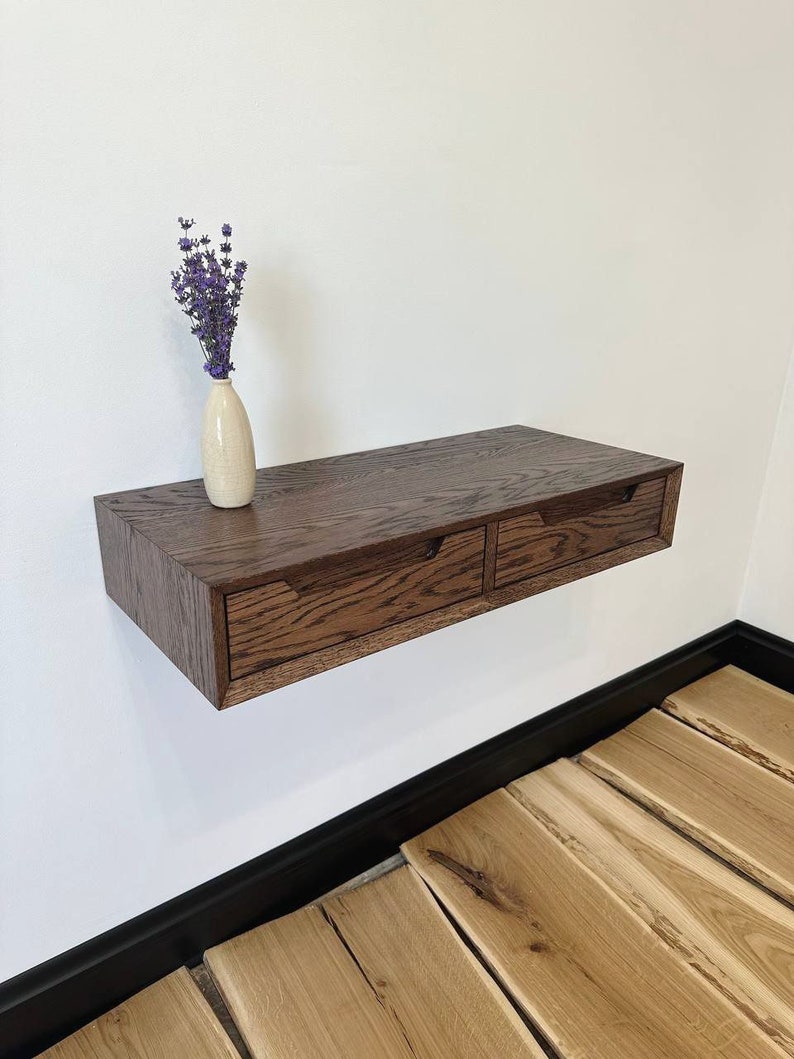May include: A dark brown wooden floating shelf with two drawers. The shelf is mounted on a white wall and has a vase of purple flowers on top.