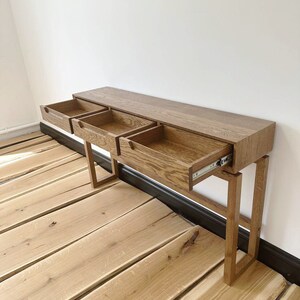 May include: A wooden console table with three drawers, all open, revealing the interior. The table has a light brown finish and is supported by four legs. The table is on a wooden floor.