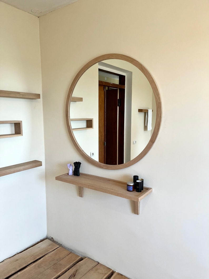 May include: A round wooden framed mirror with a shelf underneath. The shelf has two small purple figurines and three small bottles on it.