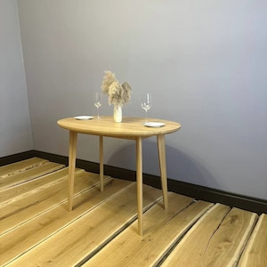 May include: A light wood oval table with a natural finish. The table has four legs and is set with two glasses and a plate. The table is in front of a light gray wall and a wooden floor.
