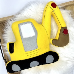 May include: A yellow plush excavator toy with black, gray, brown, and red accents. The toy features a detailed design with a cab, arm, and treads. The excavator is placed on a white fluffy surface.