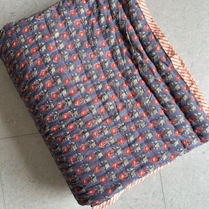 Può includere: Una coperta trapuntata piegata con un motivo floreale. La coperta presenta uno sfondo blu scuro con file di disegni floreali rossi e verdi. Il bordo è a righe arancioni e bianche.