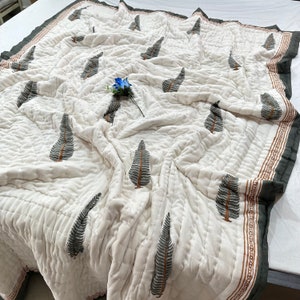 May include: White cotton quilt with a dark green tree print and a brown border. The quilt is folded and has a light blue flower on top.