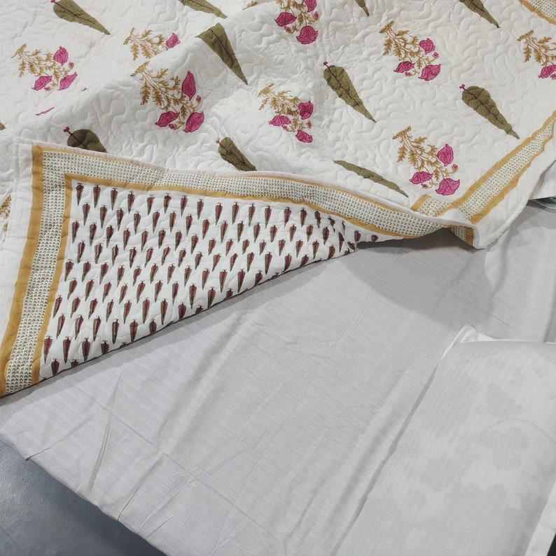 May include: A white cotton quilt with a floral print featuring pink flowers and green leaves. The quilt has a gold and white border and is folded on a white bed.