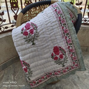 Può includere: Coperta trapuntata bianca con un design floreale rosa e verde. La coperta presenta un motivo ripetuto di fiori rosa e foglie verdi. Il bordo ha un motivo a righe verdi e bianche. La coperta è piegata su una sedia di vimini.