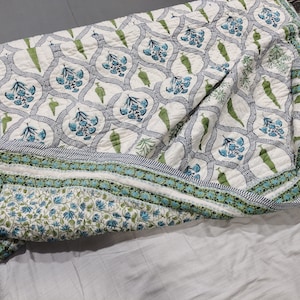 以下が含まれることがあります： 青と緑の花柄の白い綿のキルト。キルトは半分に折り畳まれ、緑と青のストライプの縁取りがあります。