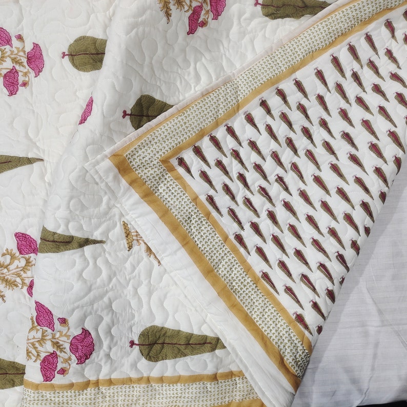 May include: A white cotton quilt with a floral print in shades of pink and green. The quilt has a gold border with a repeating pattern of small leaves. The quilt is folded in half, showing the back side, which is also white with a repeating pattern of small leaves.