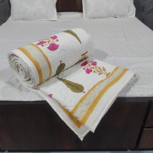 May include: White cotton quilt with a floral print in shades of pink and green. The quilt is folded and rolled up, with a yellow and white border.
