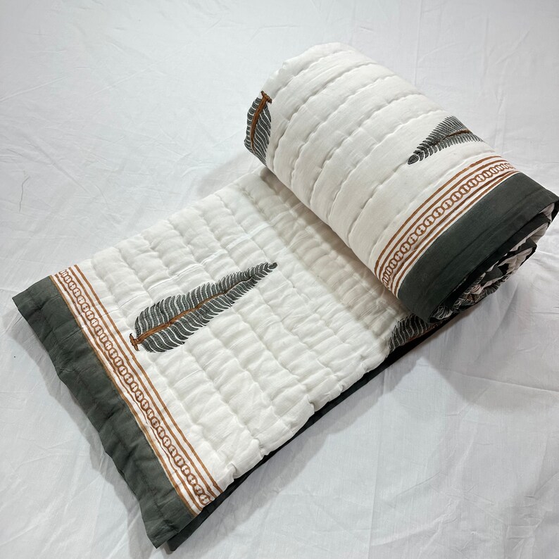 May include: White cotton quilt with a gray border and a brown and green leaf print. The quilt is folded in half and is lying on a white surface.