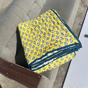 Può includere: Una trapunta gialla con un motivo floreale blu e bianco, piegata su un divano grigio. La trapunta ha un bordo blu scuro. La trapunta misura circa 152 cm per 178 cm.