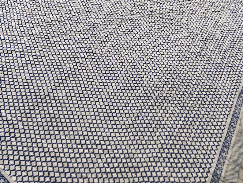 May include: A textile fabric featuring a repeating small-scale navy blue and white geometric quatrefoil pattern across the surface, with a distinct border design along the edge consisting of a thin striped band and a decorative floral motif.