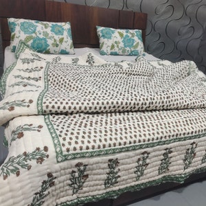 May include: A white cotton quilt with a green and brown floral print. The quilt is folded on a bed with two floral patterned pillows.
