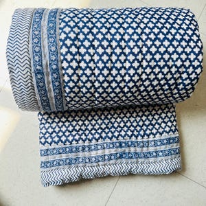 May include: A folded quilt featuring a dark blue and white cross pattern across the main body. The edges are finished with a decorative border containing a floral vine motif and a separate section with a repeating chevron zigzag pattern.