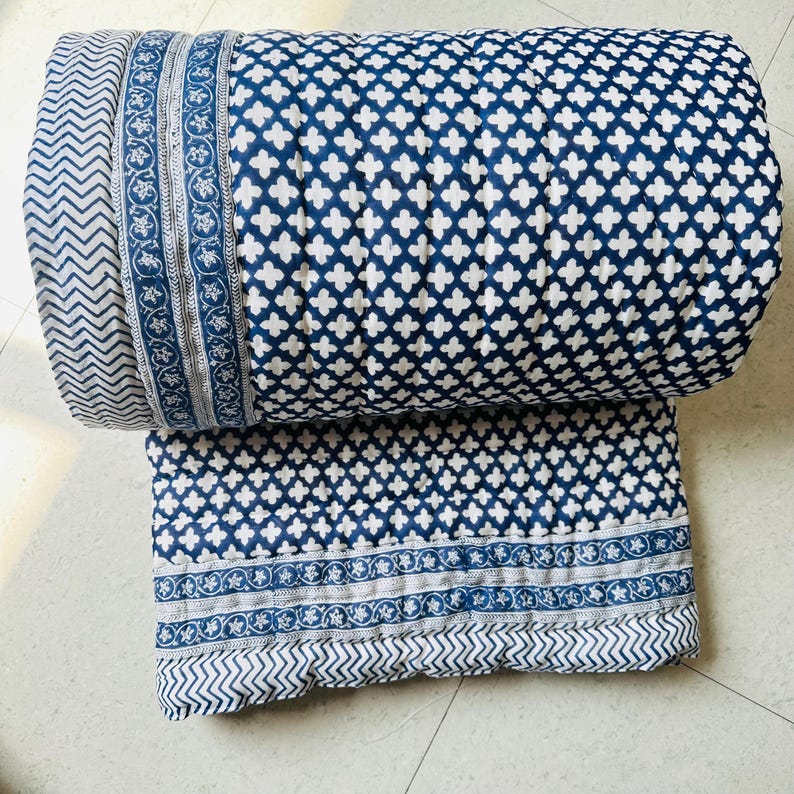 May include: A folded quilt featuring a dark blue and white cross pattern across the main body. The edges are finished with a decorative border containing a floral vine motif and a separate section with a repeating chevron zigzag pattern.