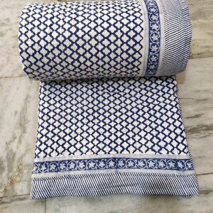 May include: A quilted textile featuring a repeating blue and white quatrefoil pattern. The fabric includes a decorative border with a floral motif and a section of thin blue and white stripes. The material is folded with a rolled top section.