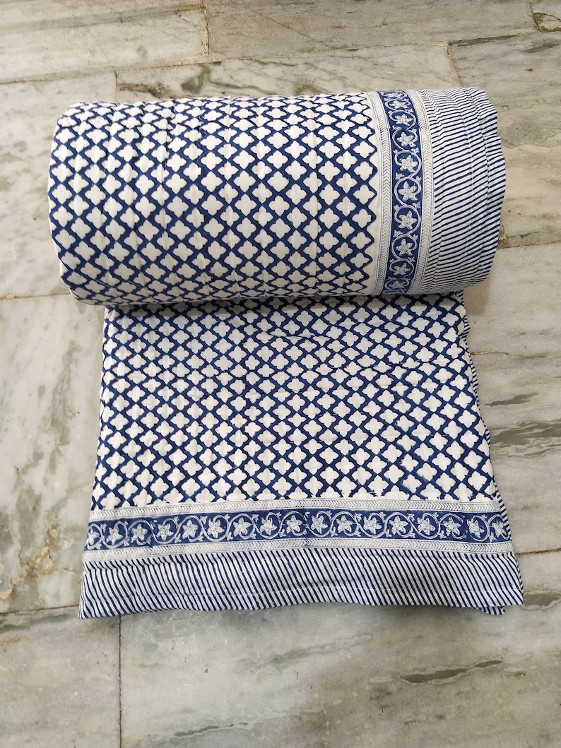 May include: A quilted textile featuring a repeating blue and white quatrefoil pattern. The fabric includes a decorative border with a floral motif and a section of thin blue and white stripes. The material is folded with a rolled top section.