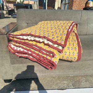 Può includere: Una trapunta gialla piegata con un motivo floreale e un bordo bordeaux è appoggiata su un divano grigio. Il design della trapunta presenta motivi floreali rosa e verdi ripetuti su uno sfondo giallo, con un lato inferiore bianco.