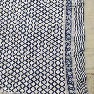 May include: A quilted textile featuring a repeating dark blue quatrefoil pattern on a white background. The border consists of a floral vine motif in blue and white, finished with an outer edge of thin, diagonal blue and white stripes.