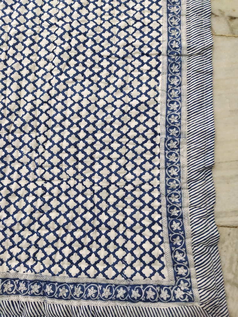 May include: A quilted textile featuring a repeating dark blue quatrefoil pattern on a white background. The border consists of a floral vine motif in blue and white, finished with an outer edge of thin, diagonal blue and white stripes.