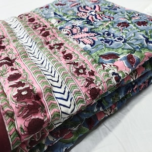 Può includere: Trapunta piegata con motivi floreali rosa, blu e bordeaux. La trapunta ha una sezione a righe con sfondo bianco e motivo a zig-zag verde e blu. I motivi floreali includono foglie e fiori.