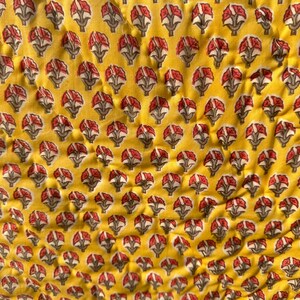 Può includere: Primo piano di un tessuto giallo con un motivo floreale ripetuto. I fiori sono rossi e bianchi con steli grigi, su uno sfondo giallo brillante. Il tessuto ha una consistenza leggermente stropicciata.