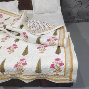 May include: White cotton quilt with a floral print in shades of pink and green. The quilt has a gold border and is folded on a bed.