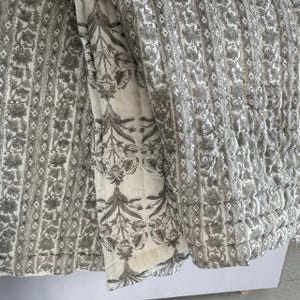 Può includere: Un trapunta di cotone bianco con un motivo floreale bianco e nero. La trapunta è composta da due pezzi di tessuto cuciti insieme, uno con un motivo ripetitivo di piccoli fiori e l'altro con un disegno floreale più grande.