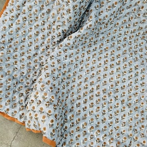 May include: A light blue quilted textile featuring a repeating pattern of small brown and white flowers. The fabric has a soft, textured appearance with visible stitching lines throughout and a solid orange border along the bottom edge.