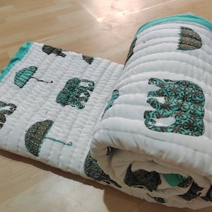 Puede incluir: Edredón de algodón blanco con un borde turquesa y un patrón repetido de elefantes y paraguas. El edredón está enrollado y el patrón es visible en el exterior del rollo.