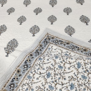 Può includere: Un trapunta di cotone bianco con un motivo floreale blu e marrone. La trapunta ha un bordo con un motivo floreale ripetitivo.