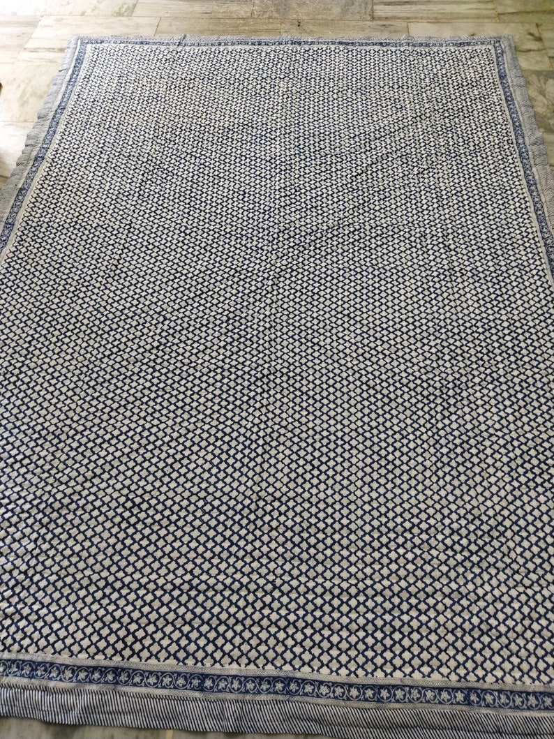 May include: A rectangular textile featuring a repeating dark blue and white diamond grid pattern across the centre. The border consists of a dark blue floral motif on a white background, finished with a thin, dark blue and white striped edge.