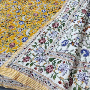 Può includere: Una trapunta a fantasia floreale. La trapunta ha uno sfondo giallo con fiori blu e rossi. Il bordo è bianco con un motivo floreale ripetuto. La trapunta è piegata, mostrando entrambi i lati del tessuto.