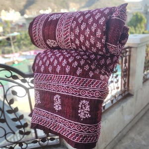 May include: A rolled-up burgundy quilt with a white floral and geometric pattern. The quilt is made of a soft material and is folded over a wrought iron railing. The background is blurred, suggesting an outdoor setting.