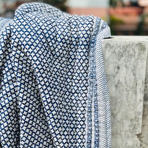 May include: A quilted textile featuring a repeating dark blue and white cross-shaped pattern. The fabric has a distinct border with a narrow stripe of small floral motifs and a wider band of blue and white chevron zig-zag lines along the edge.