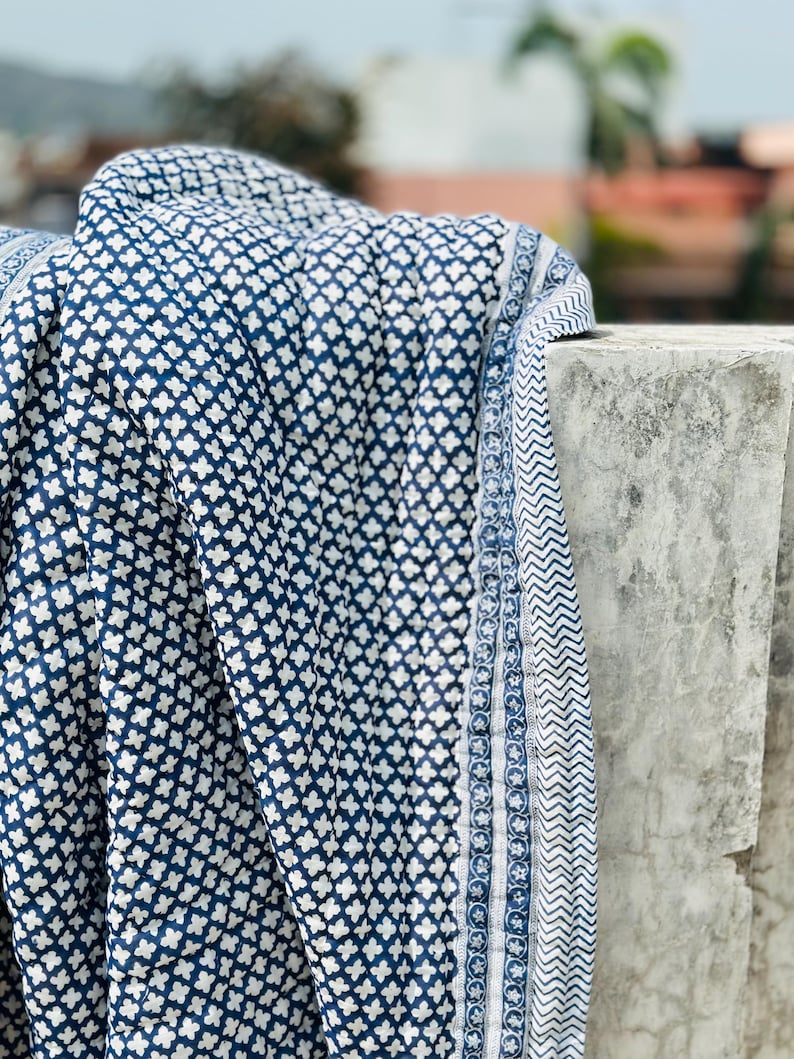 May include: A quilted textile featuring a repeating dark blue and white cross-shaped pattern. The fabric has a distinct border with a narrow stripe of small floral motifs and a wider band of blue and white chevron zig-zag lines along the edge.