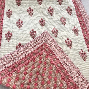 Può includere: Un trapunta di cotone bianco con una stampa floreale rosa. La trapunta ha un bordo di disegni floreali rosa e rossi. La trapunta è piegata a metà e l'angolo è visibile.