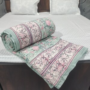 Può includere: Un piumino di cotone con motivo floreale verde e rosa con un design a righe. Il piumino è arrotolato e posizionato su un letto bianco a righe.