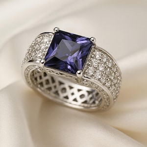 May include: A silver ring with a large, square, deep purple gemstone at the center. The band is encrusted with smaller, clear gemstones. The ring is set against a white, silky background, highlighting its elegant design. A piece of jewelry.