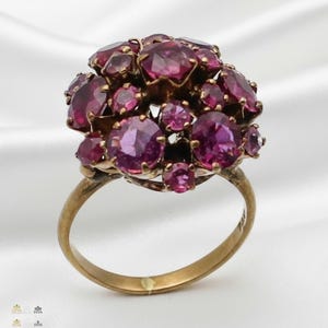 May include: A vintage gold ring with a cluster of oval and round-cut, deep pink gemstones. The ring's band is a simple, polished gold, and the gemstones are set closely together, creating a floral-like design. The ring is set against a white background.