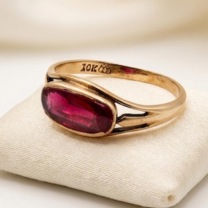 Puede incluir: Un anillo de oro con una piedra preciosa de color rojo intenso en forma ovalada montada horizontalmente. La banda está hecha de oro de 10K, con el sello 10K claramente grabado en la superficie interior. El diseño incluye un brazo dividido.